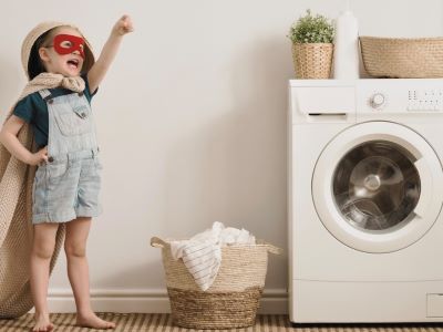 Toddler posing as a superhero with a cape and mask standing next to a washing machine and basket of laundry